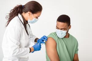 Covid-19 Vaccination. General Practitioner Vaccinating African American Male Patient Over White Studio Background, Applying Adhesive Plaster On Arm After Giving Vaccine Injection To Guy In Face Mask photo