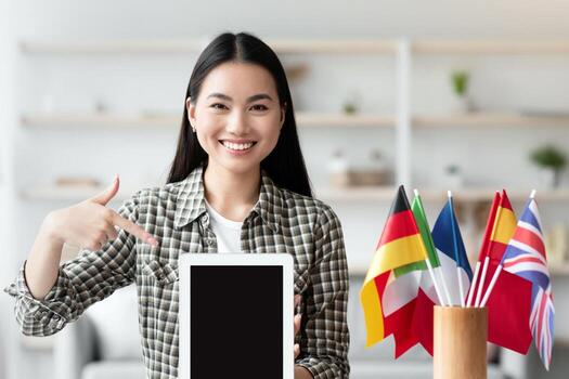 Happy asian woman pointing at pad with blank screen, sitting at worktable with flags of different countries. Cheerful young lady recommending app for foreign languages learning, showing digital tablet photo