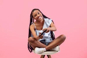 Full length of happy female gamer with joystick playing game on chair over pink studio background. Joyful young black woman with controller taking part in computer gaming competition photo