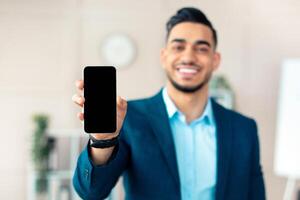 Positive Arab businessman holding smartphone with empty screen at office, mockup for mobile app or website design. Confident guy manager showing blank cellphone, copy space. Selective focus photo