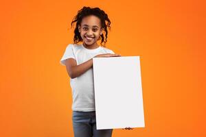 Cheerful African American Girl Holding And Showing Blank Paper Poster For Your Text Smiling To Camera Standing Isolated Over Yellow Studio Background. Kid With White Vertical Board For Advertising photo