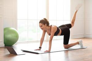 Your Personal Trainer. Female fitness instructor showing exercises while streaming, broadcasting lesson on training at home using laptop. Sport, online gym concept. Lady doing donkey kicks photo