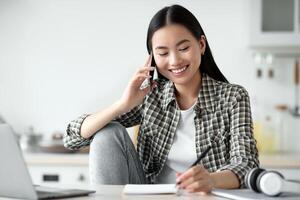 Smiling young asian woman freelancer talking on mobile phone and taking notes, working from home during isolation, copy space. Happy lady having conversation on phone, using laptop at kitchen photo
