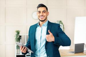 Smiling Arab businessman holding computer, showing thumb up gesture in office. Handsome guy entrepreneur recommending online services, having virtual conference with colleagues photo