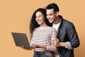 Young middle eastern couple using laptop together while standing over yellow background in studio, cheerful arab spouses shopping online or browsing new website on computer, panorama with copy space photo