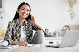 Smiling young asian woman freelancer talking on mobile phone and using laptop, working from home during isolation, looking at copy space. Happy lady having conversation on phone at kitchen photo