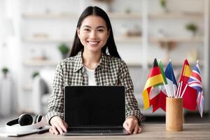Cheerful asian woman showing laptop with blank screen, sitting at worktable with flags of different countries. Smiling young lady recommending website for foreign languages learning, home interior photo