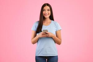 People And Technology Concept. Portrait of happy smiling young woman using smartphone standing isolated over yellow studio background, copy space. Lady chatting online, looking at camera photo
