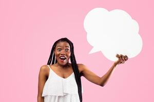Shocked Afro woman holding blank speech bubble with mockup for ad design on pink studio background. Surprised black woman with cornrows showing empty word box, free space photo