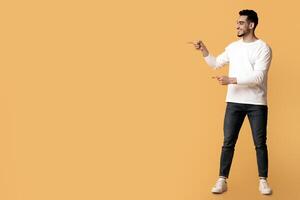 Place For Ad. Cheerful Arab Man And Woman Pointing At Copy Space In The Middle, Happy Middle Eastern Couple Recommending Something While Standing Over Yellow Studio Background, Full Length, Panorama photo