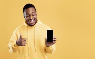 Great Mobile Application. African American Guy Showing Cellphone With Empty Screen Gesturing Thumbs Up Posing On Yellow Background In Studio. Technology And Gadgets. Mockup photo