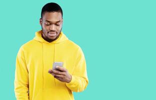 Black Young Man Using Smartphone Texting In Social Media Or Using New Mobile Application Standing Over Blue Studio Background, Wearing Yellow Hoodie. People And Gadgets, Technology Concept photo