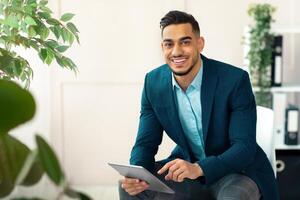 Handsome Arab businessman in formal suit using tablet pc at workplace, looking through web documents in office. Successful young entrepreneur checking email, having online business meeting photo