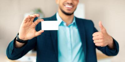 Unrecognizable Arab entrepreneur holding empty business card, gesturing thumb up at office, mockup for design. Cropped view of Eastern businessman demonstrating blank advertising card photo