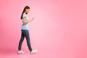 Communication, Technology And Lifestyle Concept. Full length portrait of beautiful smiling young woman using mobile phone, walking isolated over orange studio background, vertical shot photo