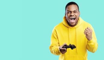 Gaming Concept. Emotional Black Guy Winning Game Posing With Joystick And Gesturing Yes In Joy Over Blue Studio Background. Millennial Man Celebrating Victory Playing Videogame. Panorama photo