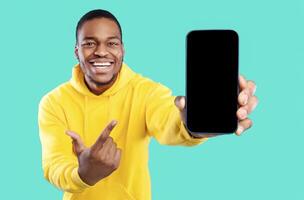 Cheerful African American Man Showing Cellphone With Blank Screen Advertising Great Application And Pointing Finger At Smartphone Posing Over Blue Studio Background. Check This New App. Mockup photo