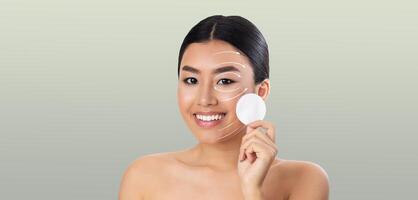 Asian woman with a radiant smile engages in her skin care routine, gently applying a product to her face. She is isolated against a calming background, focusing on self-care and beauty. photo