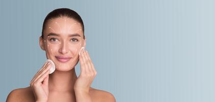 A woman engages in her skincare routine, gently cleansing her face with a cotton pad. She smiles, embracing a moment of self-care in a serene, isolated setting with a soft blue backdrop. photo