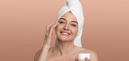 A woman engages in her skincare routine, applying cream on her face with a smile. She is wrapped in a towel, surrounded by a soft, pastel-colored background, promoting relaxation and self-care. photo