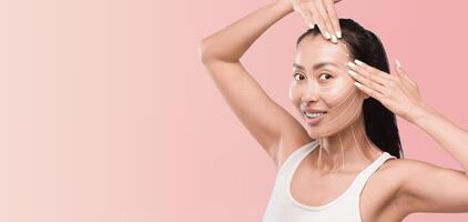 Asian woman is applying a facial mask as part of her skincare routine, surrounded by a calm, pink backdrop. The setting promotes relaxation and beauty, highlighting the pampering experience. photo