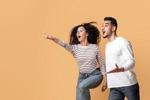 Look There. Amazed Arab Couple Pointing Aside While Jumping On Yellow Background, Excited Young Man And Woman Demonstrating Copy Space For Design Or Advertisement, Full Length photo
