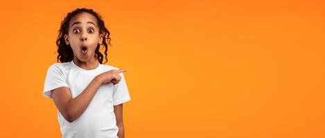 Wow, Unbelievable Offer Concept. Portrait Of Surprised African American Girl Pointing Finger Aside At Free Copy Blank Space For Text Standing On Yellow Orange Studio Wall. Look There, Panorama, Banner photo
