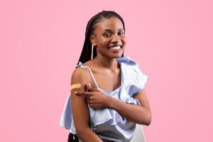 Successful covid vaccination concept. Black lady with afro bunches pointing at adhesive plaster om her arm after coronavirus immunization, pink studio background photo