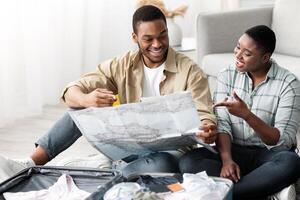 Travel Destination Choice And Navigation. Black Spouses Holding Touristic Map Choosing Country For Next Vacation Trip Together Sitting With Suitcase On Floor At Home. Tourism And Traveling Concept photo