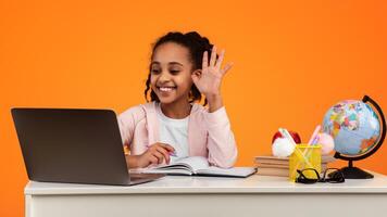 Remote Learning Concept. Portrait of positive African American schoolgirl sitting at desk, making call using laptop computer. Smiling young black girl waving hand while having virtual class photo