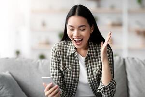 Emotional asian woman using smartphone at home, looking at gadget screen, screaming and raising hand up, reading online advert about sales or nice offer, gambling, sitting on couch, copy space photo