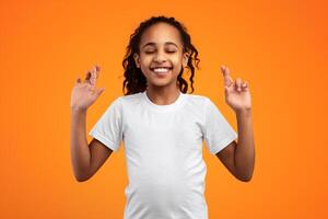 Making Cherished Wish. Positive Black Girl Crossing Fingers And Smiling, Begging For Luck, Praying For Miracle, Hoping For Good Things To Happen, Standing With Closed Eys Over Yellow Studio Background photo