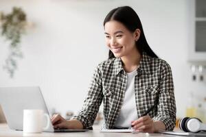 Smiling young asian woman student using laptop for studying at home, taking notes and typing on computer keyboard, copy space. Cheerful lady looking for job online, kitchen interior photo