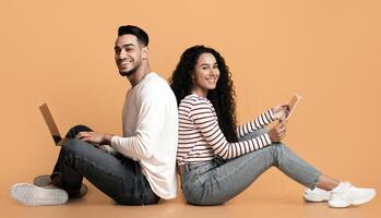 Cheerful arab couple sitting back to back with laptop and digital tablet, happy middle eastern man and woman using different gadgets while relaxing over yellow background in studio, panorama photo