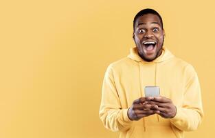 Wow News. Excited African American Guy Holding Smartphone Looking At Camera In Excitement Posing In Studio On Yellow Background. Great Mobile Application Ad Concept. Panorama photo
