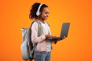 Portrait of positive black female pupil holding and using laptop computer, wearing wireless headphones and backpack, typing on keyboard, doing homework or having call with teacher, yellow studio photo