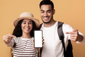 Travel Booking Online. Happy Arab Travellers Couple Pointing At Smartphone With Blank Screen, Middle Eastern Spouses Recommending Mobile App For Tour Search, Standing On Yellow Background, Mockup photo