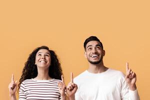 Check This. Joyful Young Arab Couple Pointing Up At Copy Space Above Their Heads, Cheerful Middle Eastern Man And Woman Showing Free Place On Yellow Background For Your Offer Or Advertisement photo