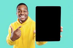 Black Guy Showing Digital Tablet With Blank Screen Recommending New App Or Great Website, Posing Pointing Finger Over Blue Background In Studio. Selective Focus, Mockup photo