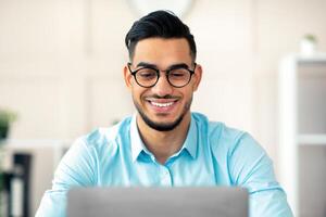 Young Arab businessman with laptop working online, making presentation at modern office. Guy company employee using computer for remote job, communication, business education photo