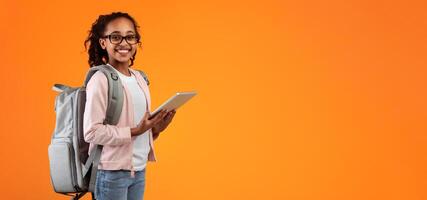Online Classes Concept. Smiling teenage pupil wearing glasses and backpack holding and using digital tablet for studying standing on yellow orange studio wall posing at camera, free copy space photo