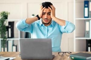 Desperate Arab businessman having too much work, making mistake, cannot meet deadline, suffering from headache at desk in office. Company employee feeling stressed from working with laptop overtime photo