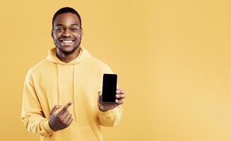 Check This Mobile Application. Black Man Showing Phone Pointing Finger At Blank Screen Smiling To Camera On Yellow Studio Background. People And Gadgets. New App For Your Cellphone. Mockup photo