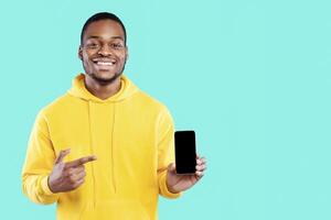 Mobile Application. Smiling African American Guy Showing Phone With Empty Screen For New App Advertisement Standing In Studio On Blue Background. People And Gadgets. Mockup photo