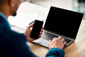 Cropped view of Arab businessman using laptop computer and smartphone with mockup for app or website design, closeup. Unrecognizable guy CEO working with modern gadgets, space for text photo