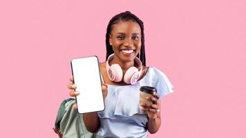 Happy black female student with backpack, headphones and coffee showing smartphone with empty screen on pink studio background, mockup for app or website design. Back to school concept photo