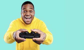Excited African American Gamer Guy Playing Game Posing With Joystick Over Blue Studio Background, Smiling To Camera. Gaming Concept. New Videogame Advertisement photo