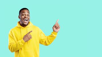 Look There. Excited African American Guy Pointing Fingers Aside Showing Blank Space For Text Posing Over Blue Studio Background, Smiling To Camera. Check This Offer Concept photo