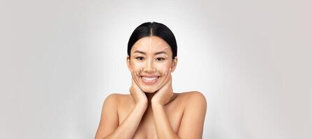 Asian woman is happily engaging in her skincare routine, gently massaging her face with both hands. She is isolated against a plain background, creating a serene atmosphere for pampering. photo