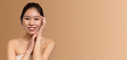 Asian woman enjoys her skin care routine, gently applying products to her face. She radiates happiness and calmness in a soothing, isolated setting with an empty background. photo